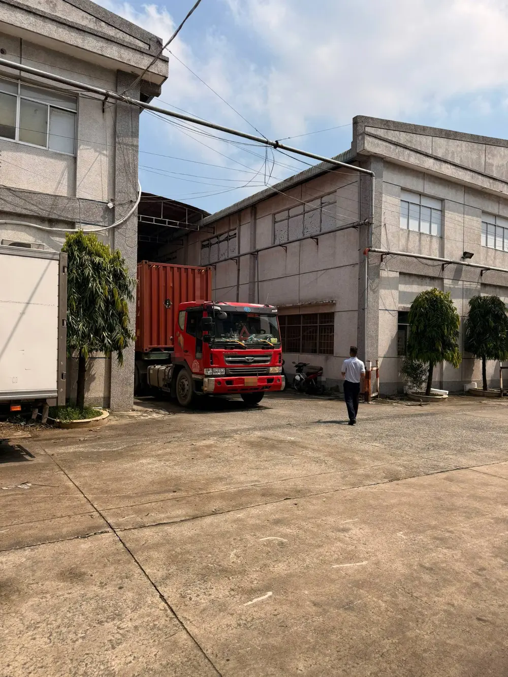Xe đầu kéo container màu đỏ đỗ ở lối vào nhà kho, một người đi bộ trên sân bê tông