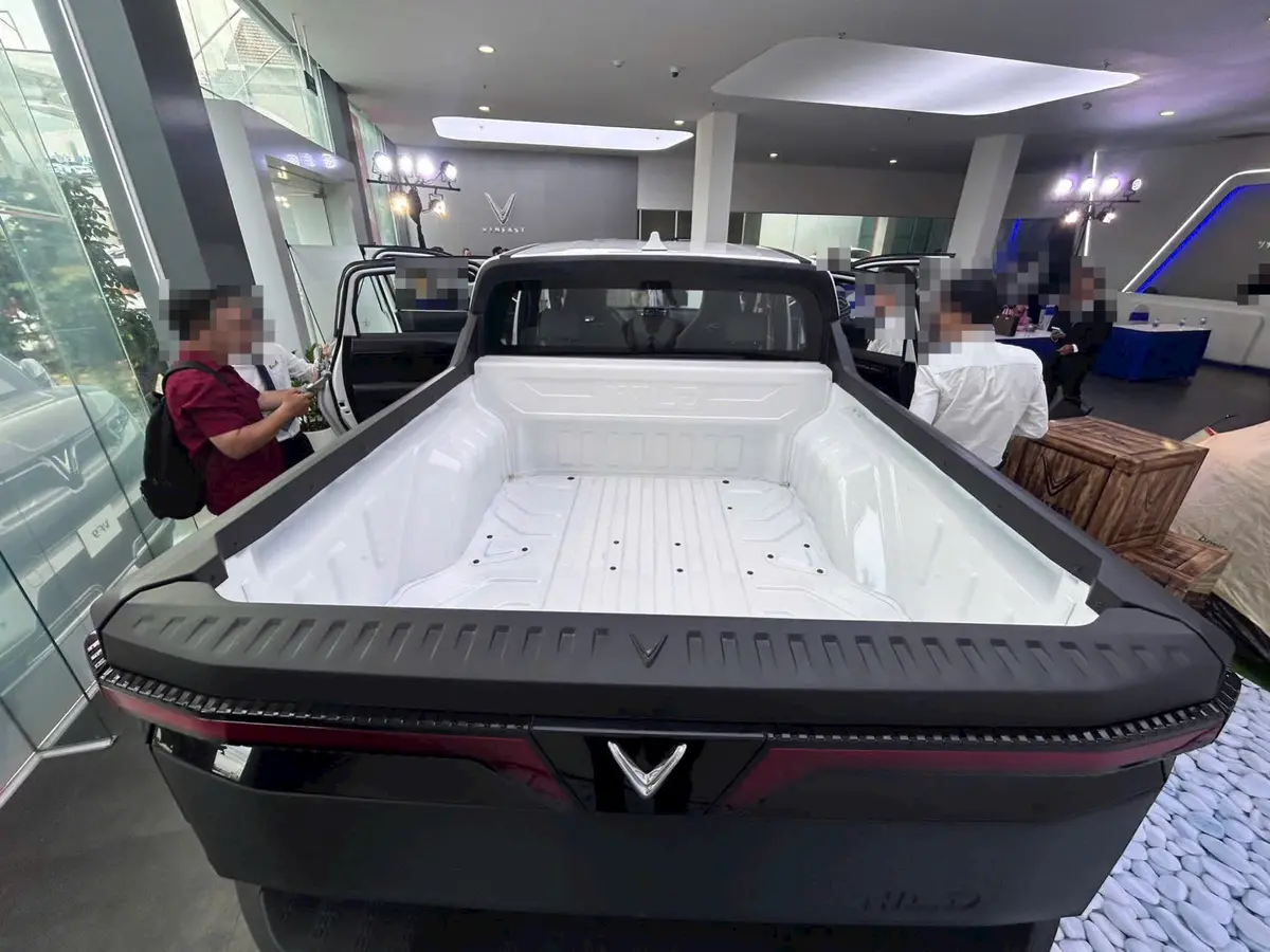 Thùng xe lót nhựa trắng của VinFast VF Wild nhìn từ phía sau, khách và nhân viên đứng quanh xe trong showroom; mặt người đã được làm mờ